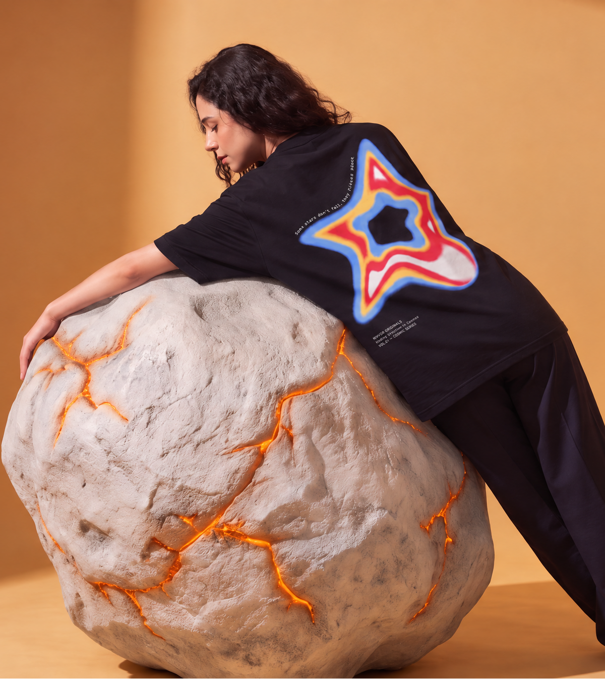 A model wearing a black oversized T-shirt with a large colorful star graphic on the back, posed next to a large rock prop in a warm studio setting. A floating product cutout of the same shirt appears on the right, showing the back graphic and small text details.